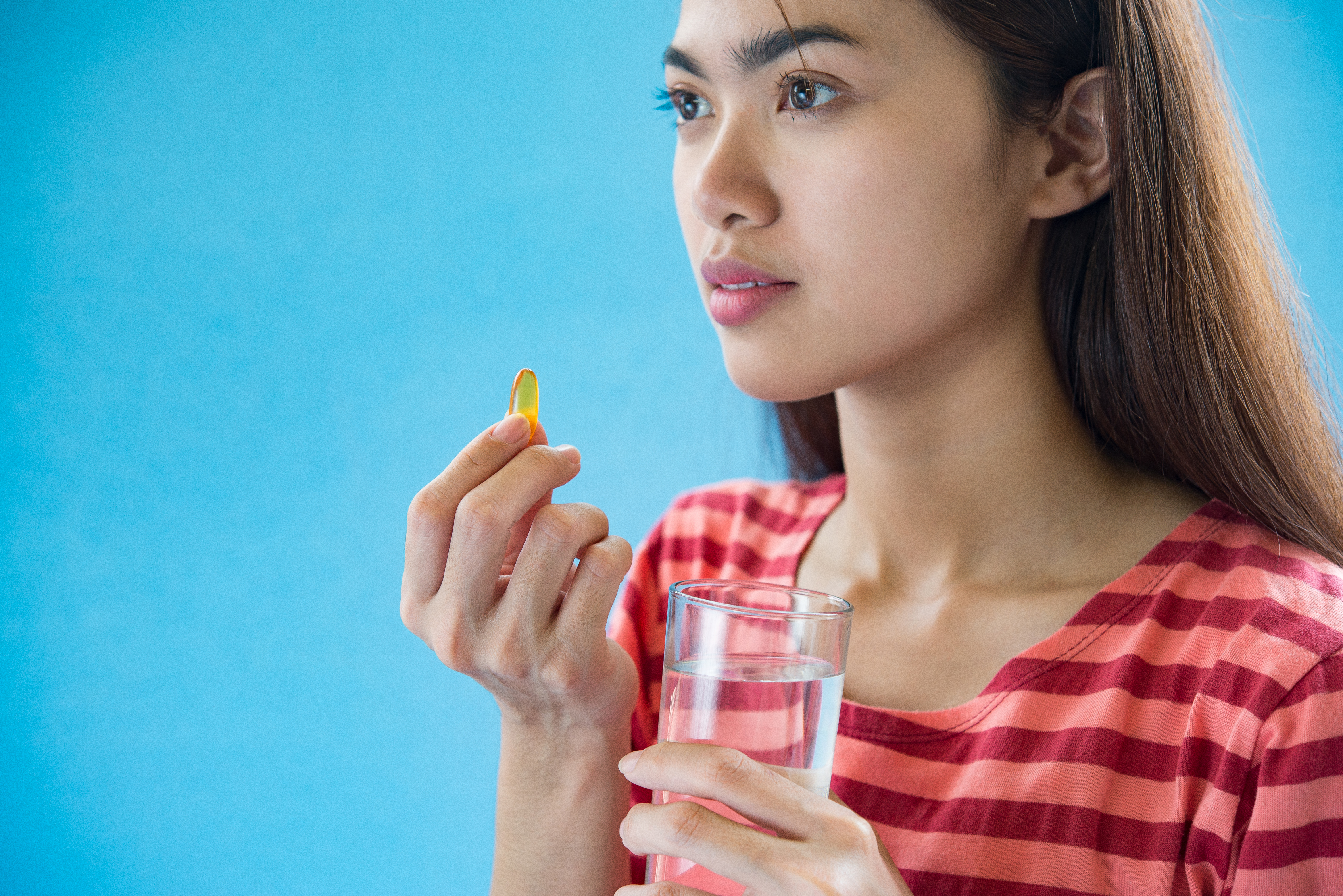 mulher de cabelos lisos e pretos parada na frente de um fundo azul tomando suas vitaminas